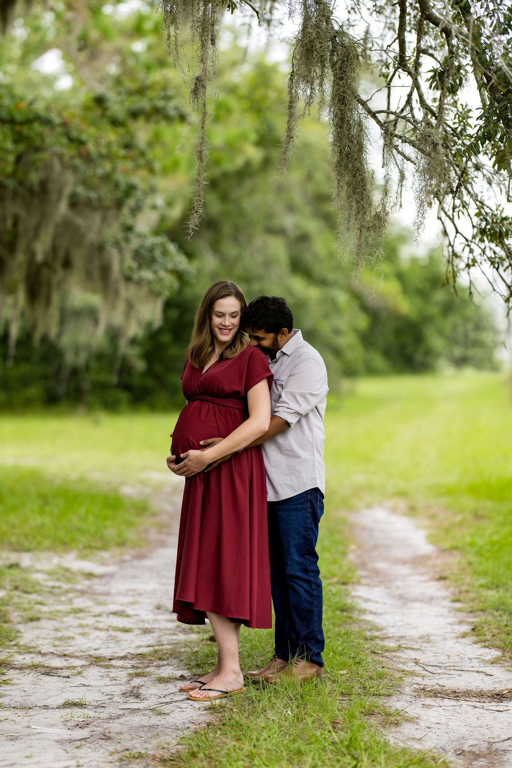Pregnant woman in red dress embraced by partner on scenic path with Spanish moss trees during maternity photoshoot.