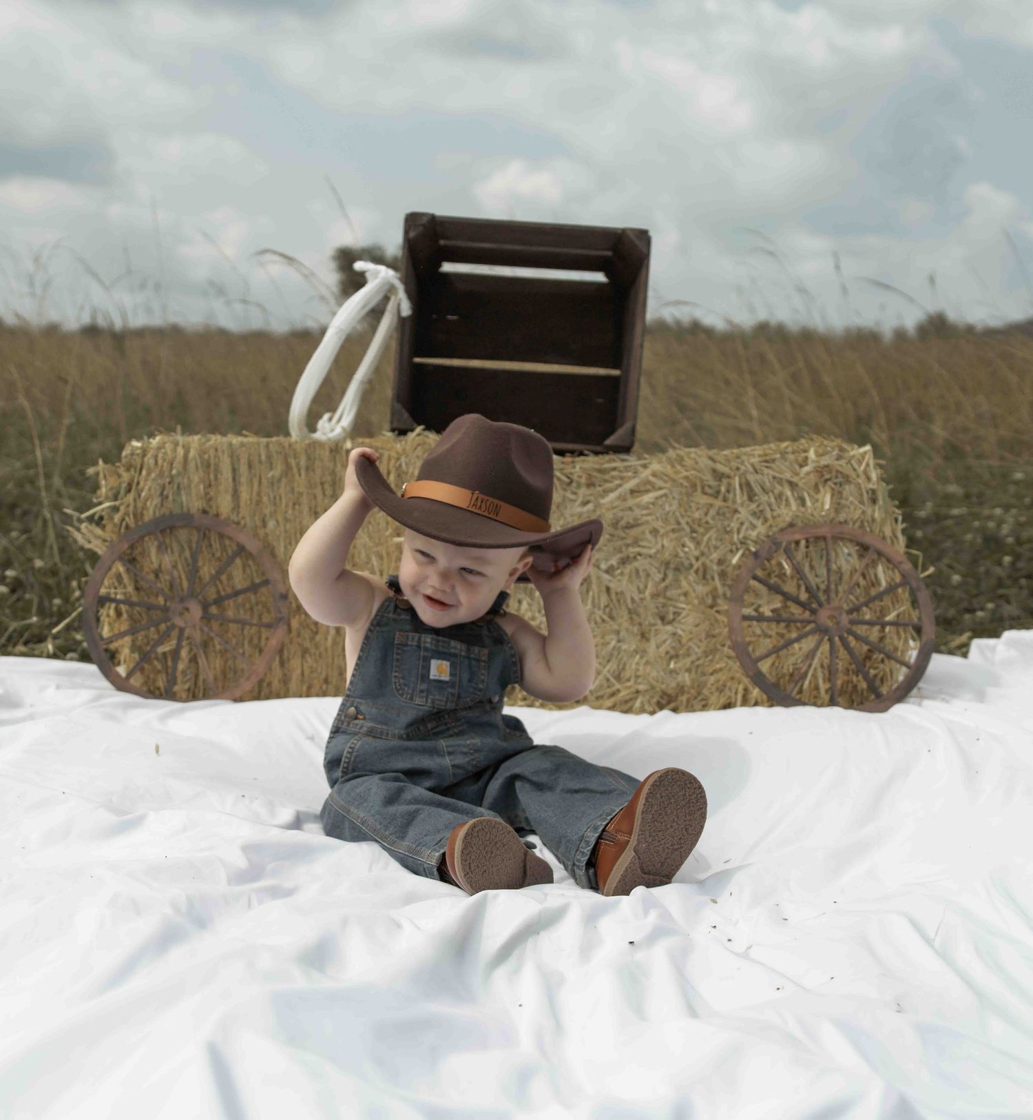 A baby in denim overalls and cowboy hat sits on white cloth near hay bales and wagon wheels in a field.