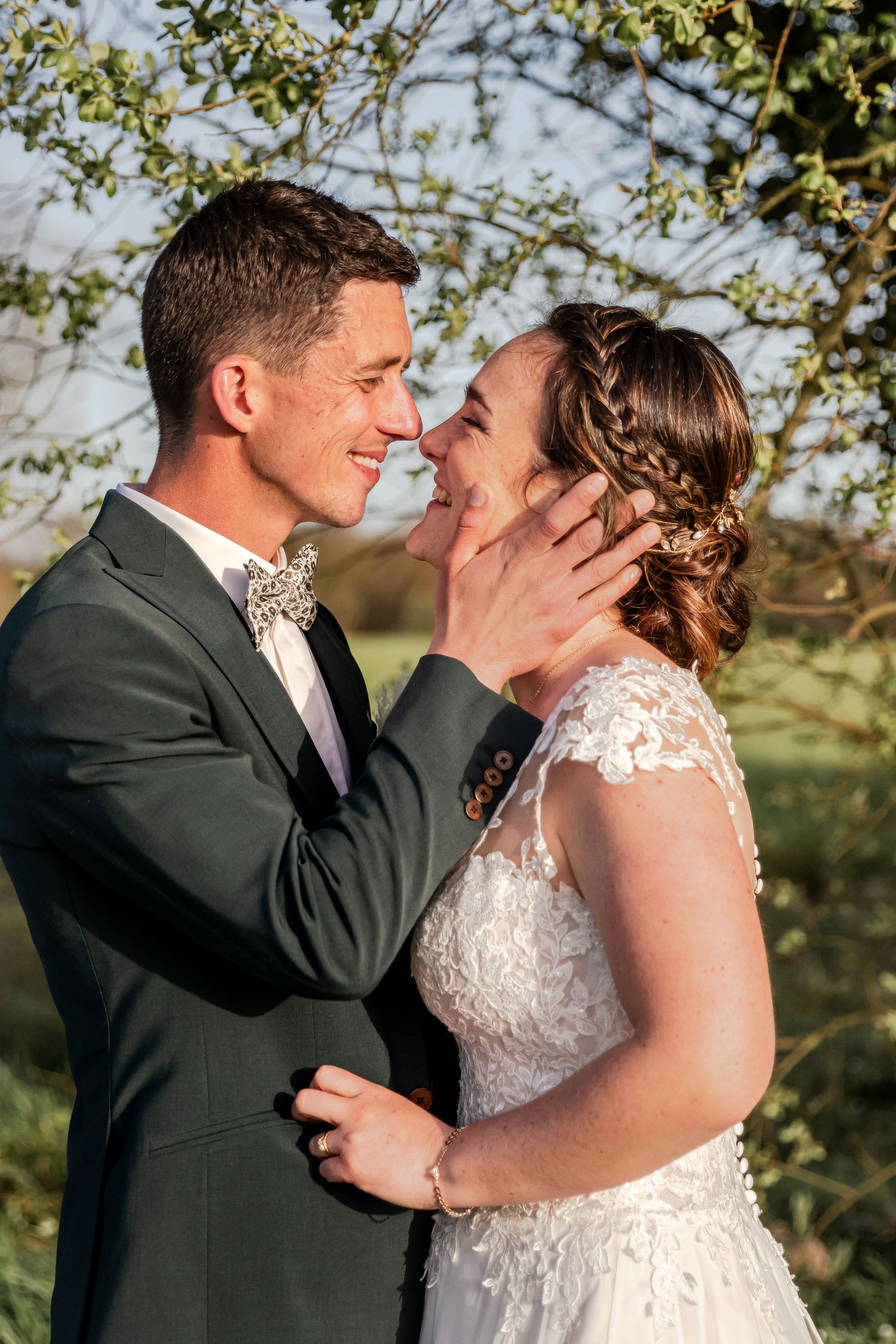 A groom in a gray suit tenderly holds his bride's face as they share a romantic moment outdoors on their wedding day.