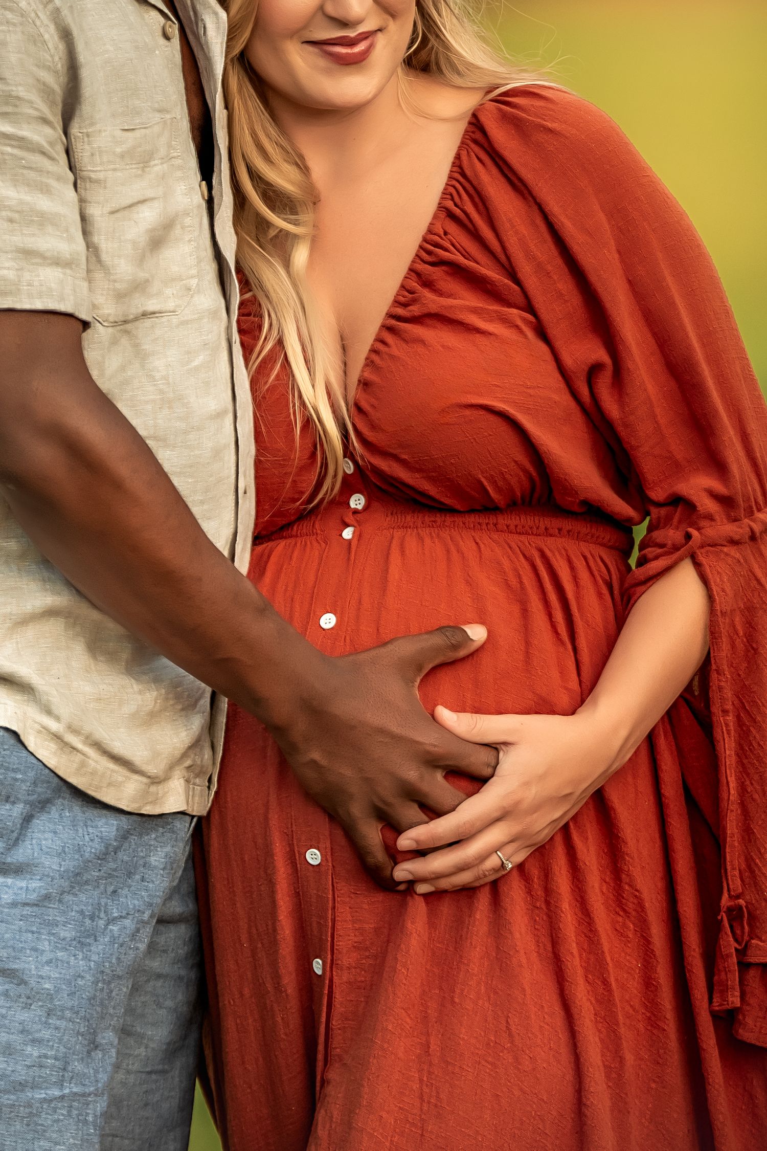 A pregnant woman in a rust-colored dress with her partner tenderly cradling her baby bump outdoors.