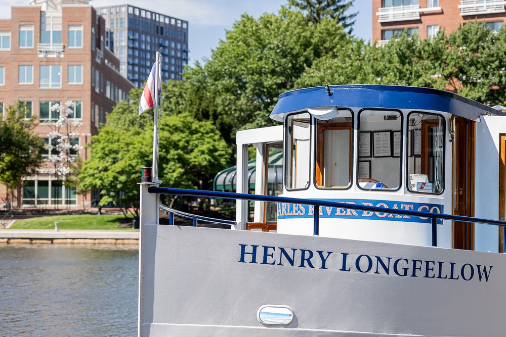 The Henry Longfellow riverboat docked on a calm waterway with city buildings and green trees in the background.