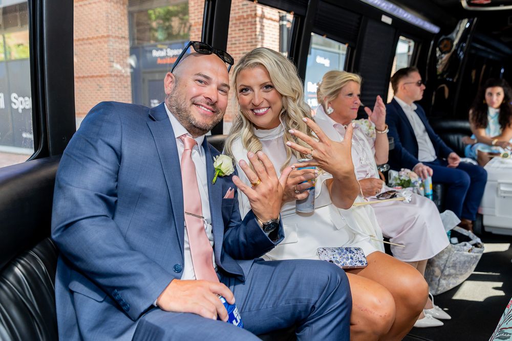 Newlywed couple smiling and showing off wedding rings inside a party bus, dressed in blue suit and white dress.
