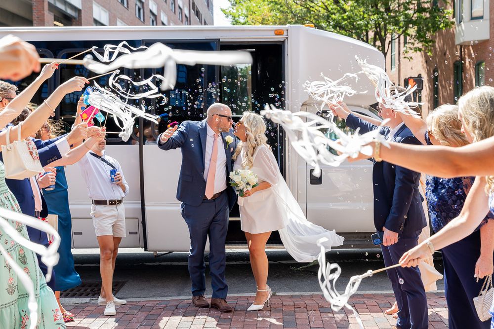 Newlyweds exit through a streamer tunnel of cheering guests in front of a white party bus on a sunny day.