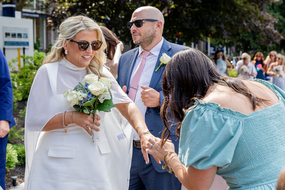Bride in white cape holding blue bouquet greeted by guests at outdoor wedding ceremony on sunny day.