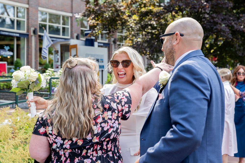 Bride in white dress laughing with guests in floral dress and blue suit at outdoor wedding celebration.