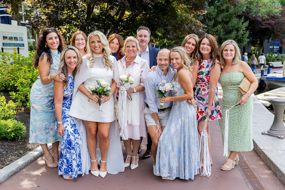 A joyful wedding group photo featuring a bride in white and guests in colorful dresses posing outdoors on a sunny day.