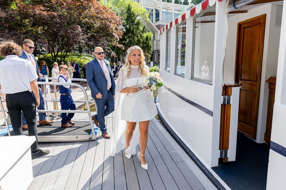 Bride in white mini dress holding bouquet walks on boat deck with guests in sunny outdoor wedding.