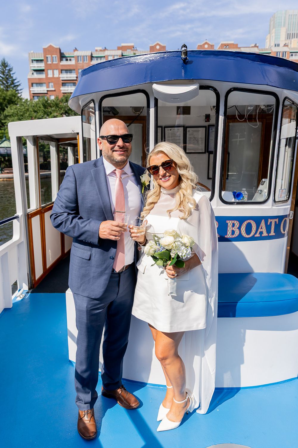 A newlywed couple poses on a blue boat deck, bride in white mini dress with bouquet, groom in blue suit holding champagne.