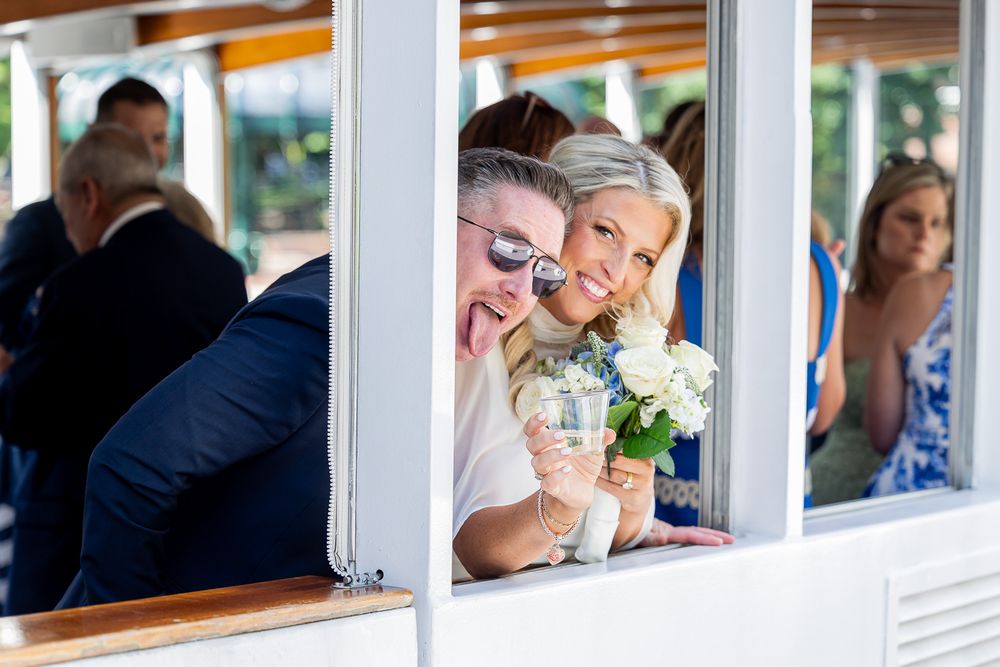 Bride and groom smiling together on a boat, bride holding white flowers, guests visible in background.