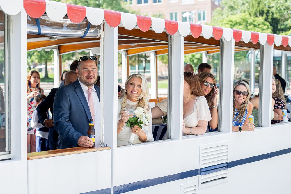 Wedding guests celebrate on a decorated boat cruise, with bride holding flowers and guests enjoying drinks on a sunny day.