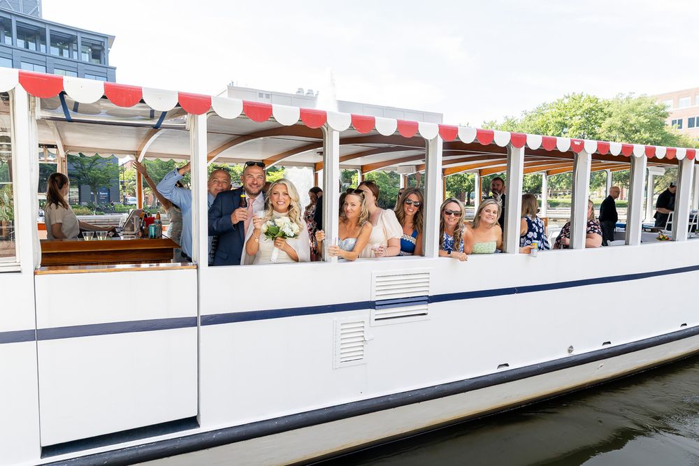 Wedding party celebrating on a decorated white canal boat with red and white awning on a sunny day.