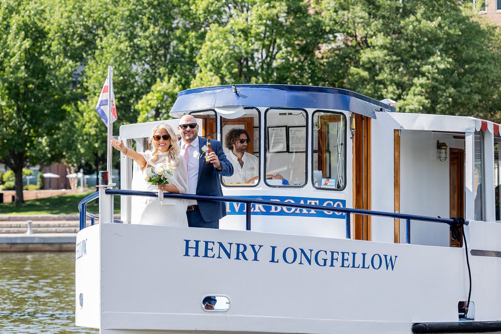Bride and groom celebrate on the Henry Longfellow boat, waving joyfully on a sunny day near lush green trees.