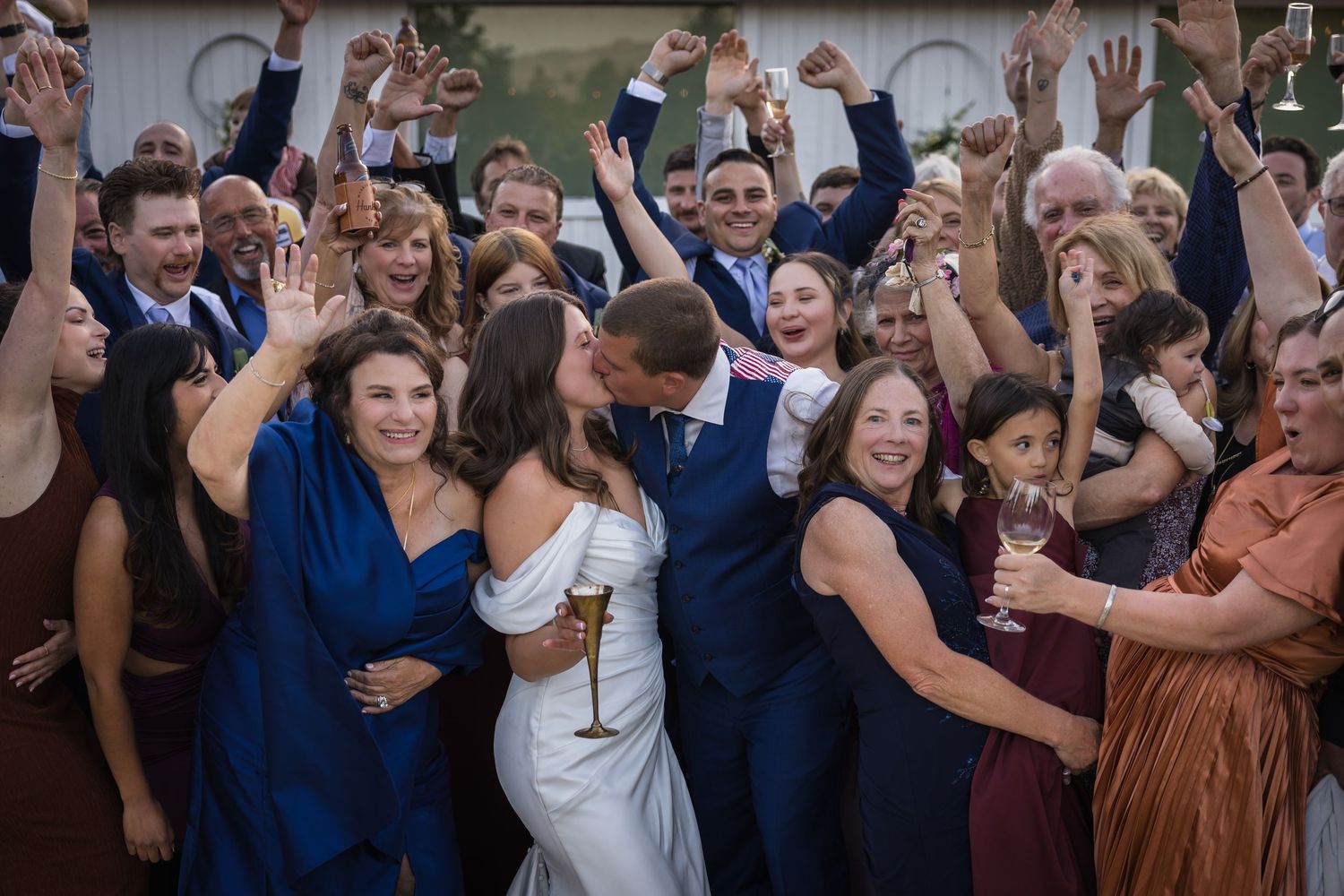 Joyful wedding couple kissing surrounded by cheering guests raising champagne glasses in celebration.