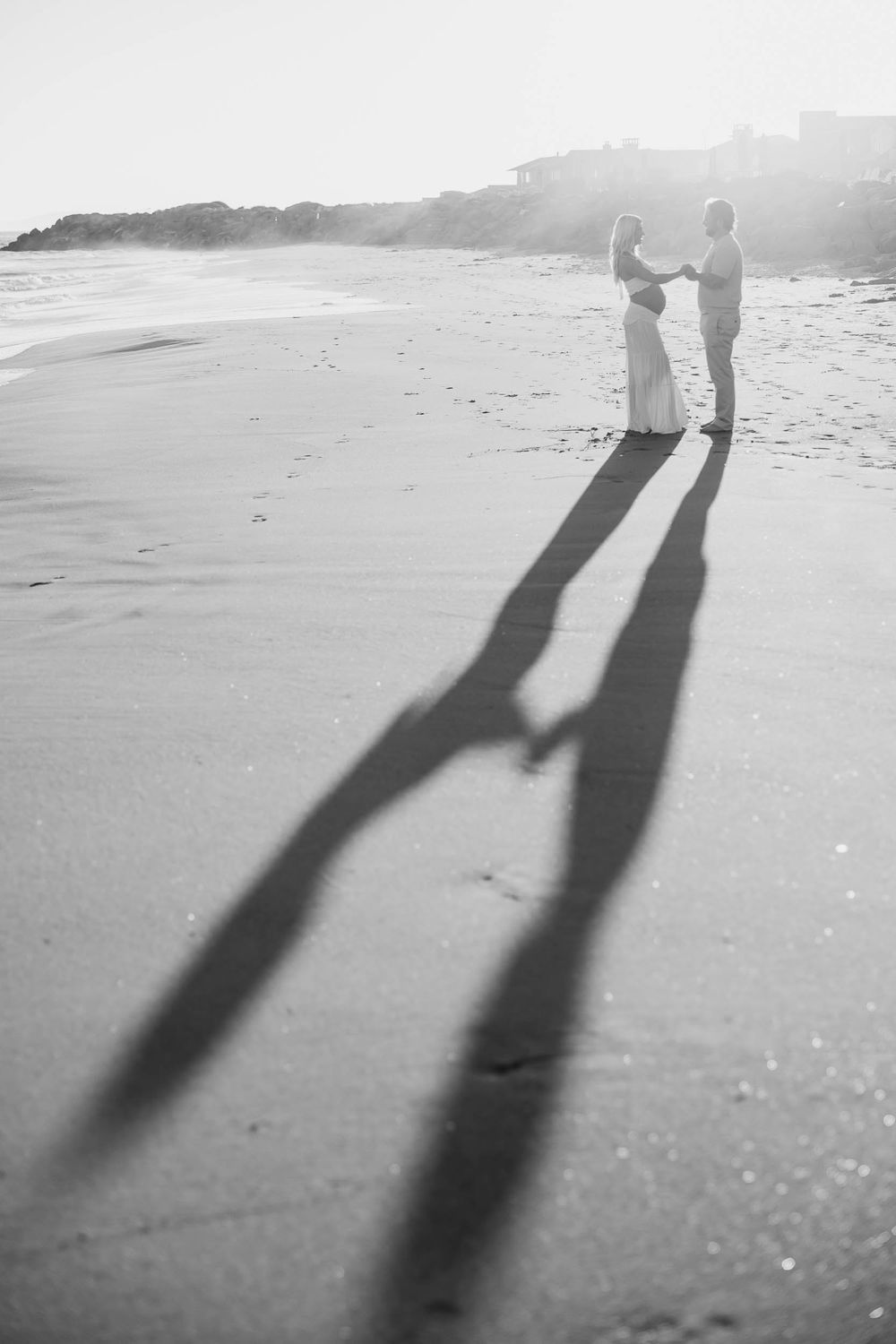 Black and white beach photo of couple holding hands at sunset, their long romantic shadows stretching across the sand.
