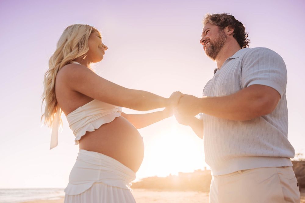 Pregnant woman in white crop top holds hands with bearded man on sunny beach, maternity photo shoot.