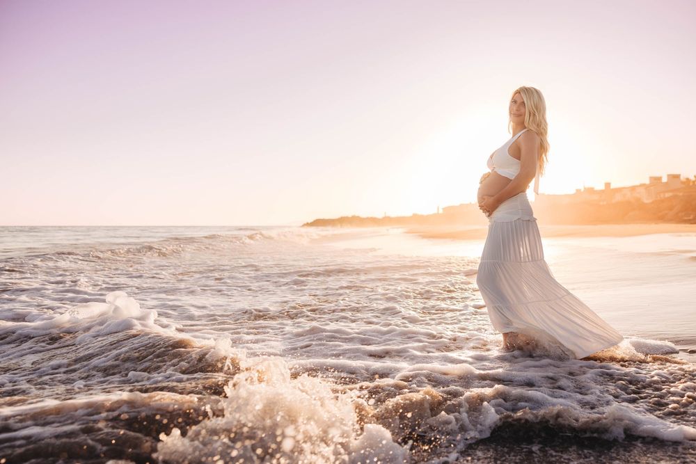 Pregnant woman in white dress standing in ocean waves at sunset on a beautiful beach.