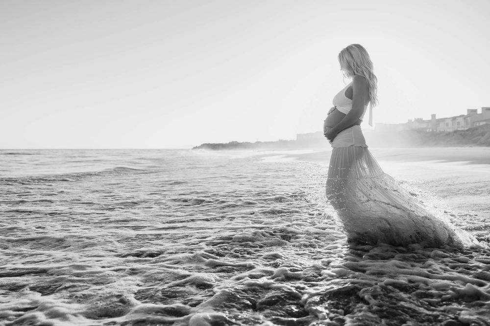 Black and white photo of a pregnant woman in a lace gown standing in ocean waves at sunset on a beach.