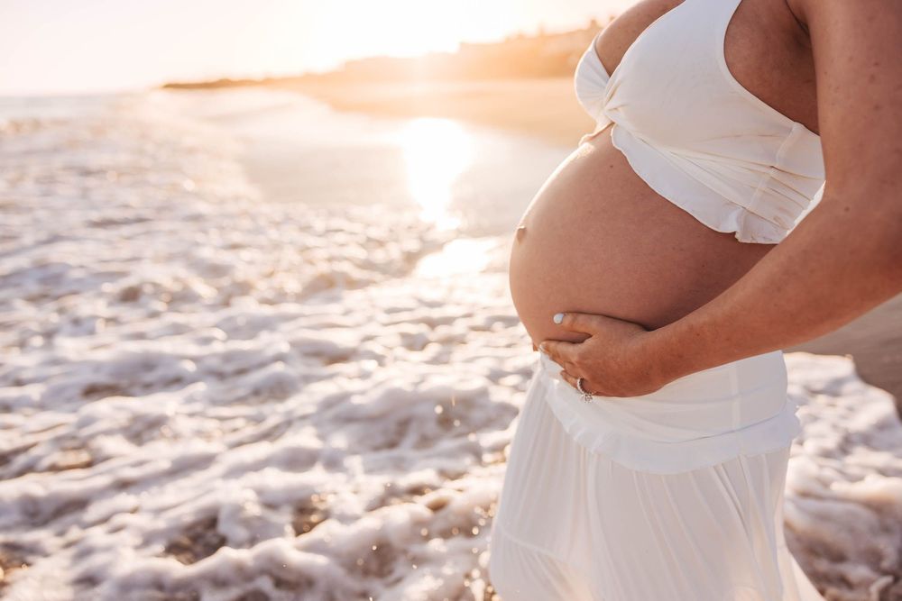 Close-up of pregnant woman in white bikini top cradling baby bump on sunny beach.