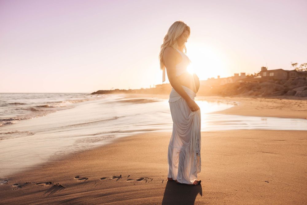 Pregnant woman in white lace dress standing on sandy beach during glowing purple sunset.