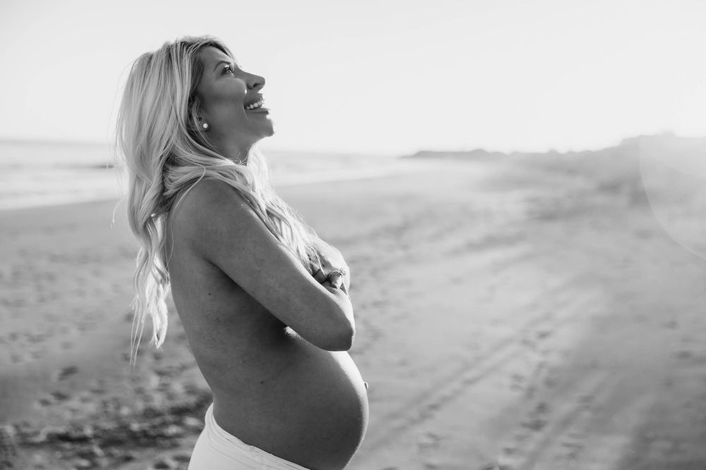 Black and white photo of smiling pregnant woman embracing baby bump on beach at sunset.