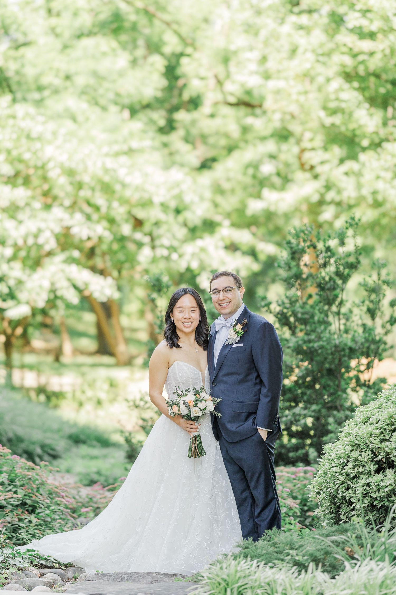 Bride and groom smiling together in a lush green garden, bride holding a floral bouquet in a strapless lace gown.