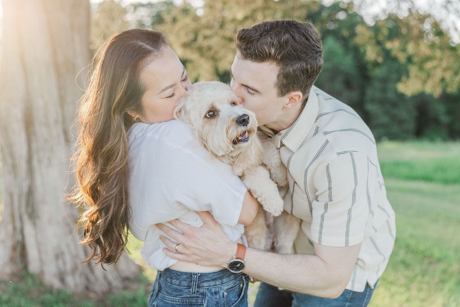 Couple sharing a kiss on their fluffy white dog during a golden hour outdoor engagement photo session.