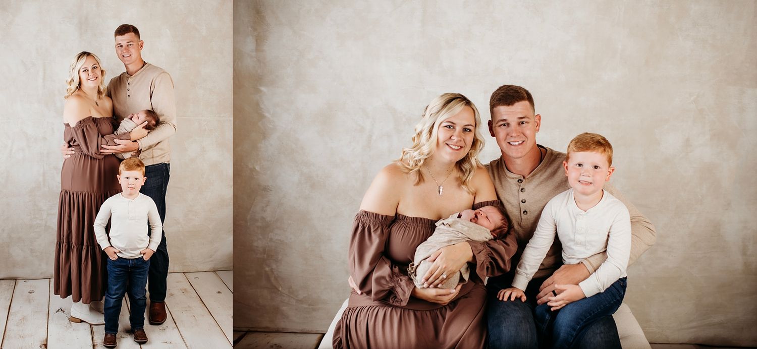 A family of four poses in a studio, parents holding a newborn while a toddler stands beside them in warm neutral tones.