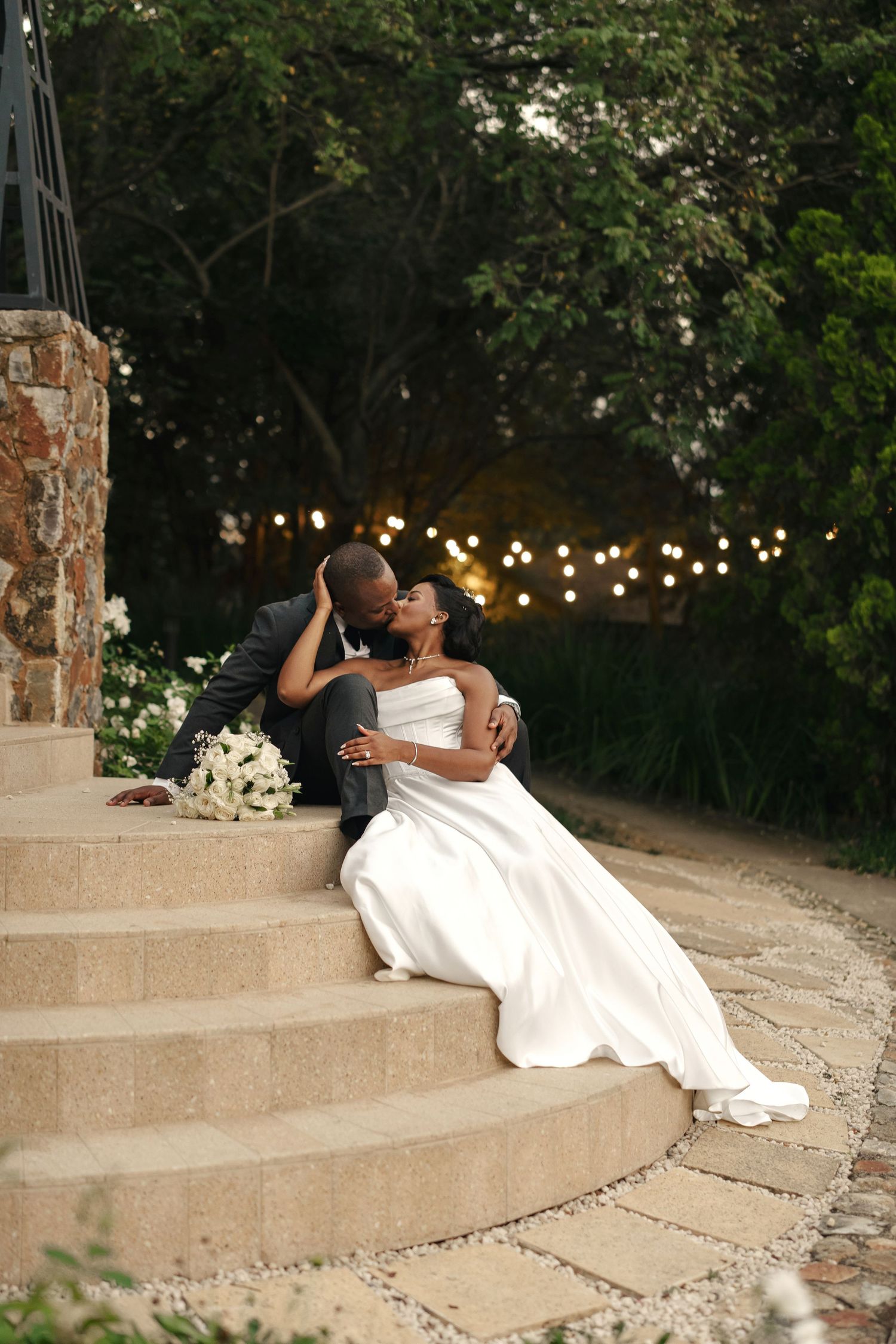 Newlywed couple sharing a romantic kiss on stone steps, bride in white gown with bouquet, groom in suit, garden lights.