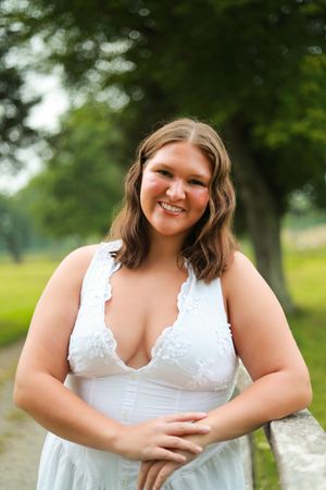Smiling young woman in white lace dress leans on fence in green park setting.