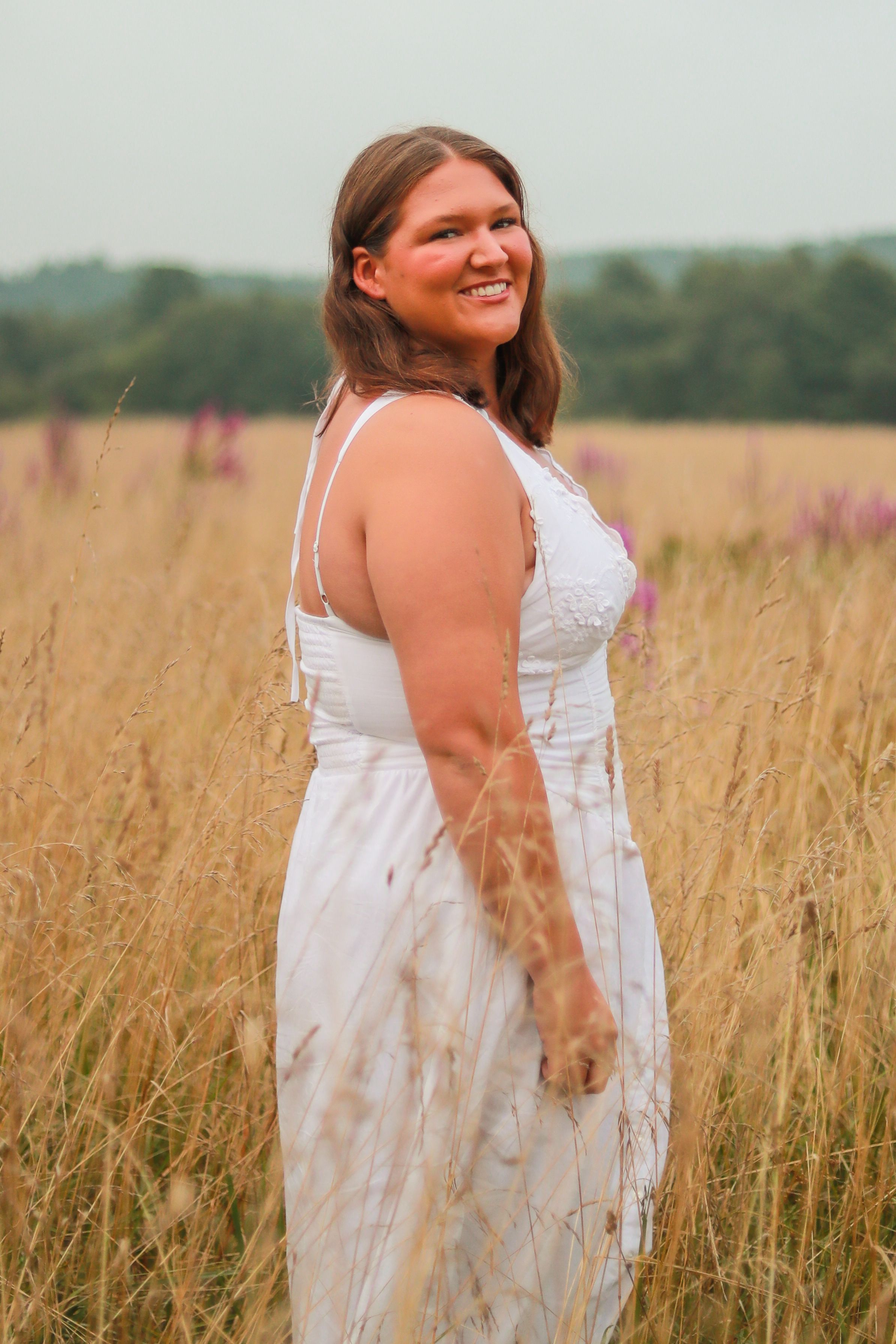 Smiling woman in white sundress stands in golden field with wildflowers in background.