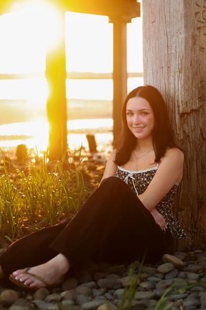 Young woman with dark hair sitting against a wooden post at sunset by the water, smiling warmly.