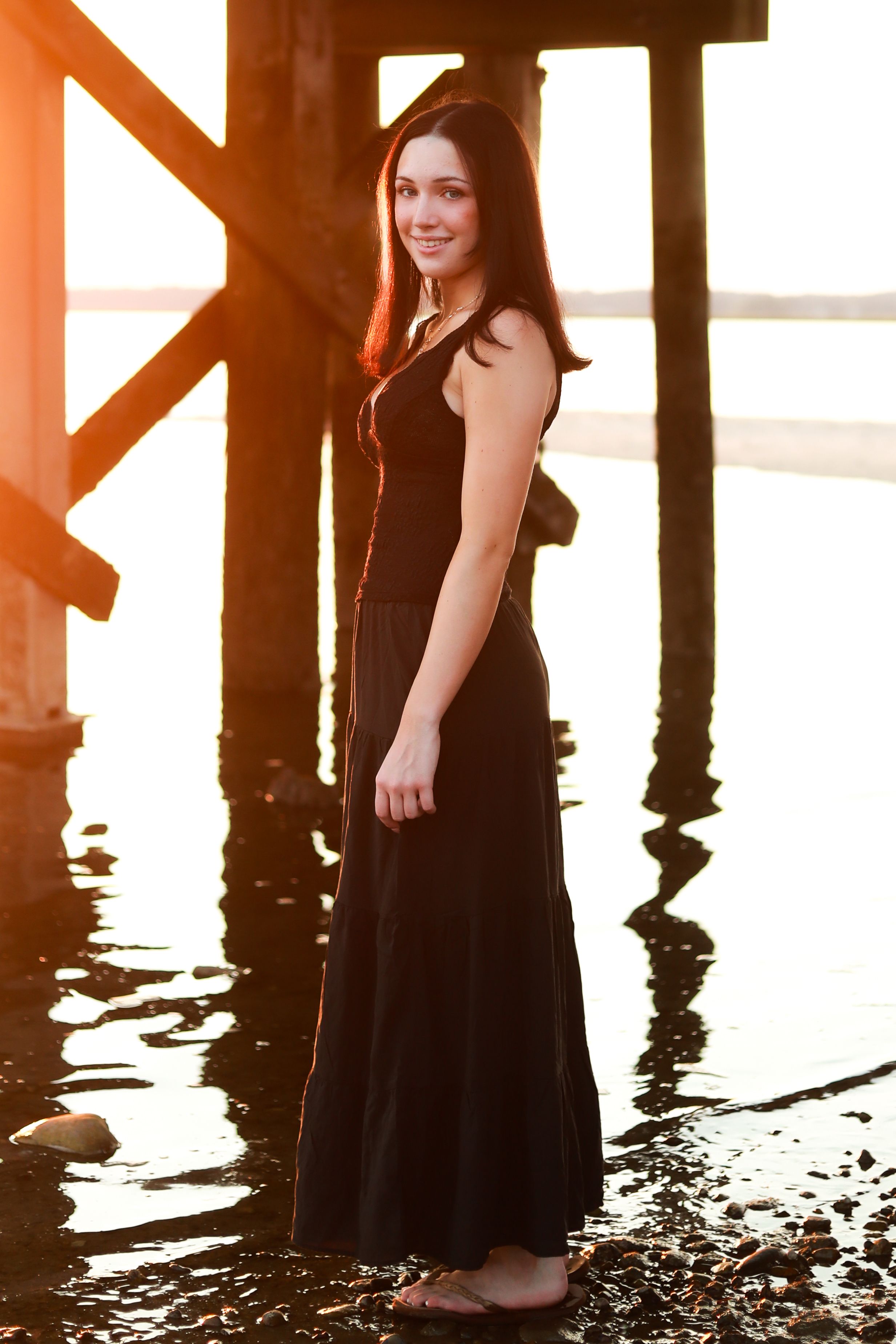 Young woman in black maxi dress standing under pier at sunset with warm golden light reflecting on water.