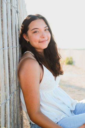Young woman with long dark hair smiles while leaning against a wooden fence outdoors in warm sunlight, wearing a white top.