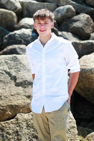Smiling teenage boy in white shirt and khaki pants posing confidently in front of large rocks outdoors.