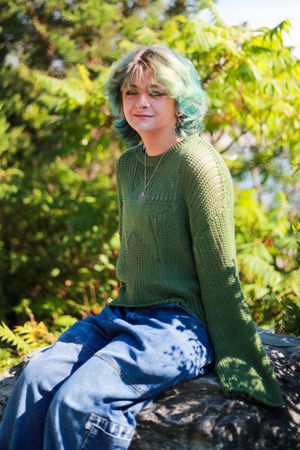A young person with green hair sits on rocks wearing a green knit sweater and wide-leg jeans, surrounded by lush foliage.