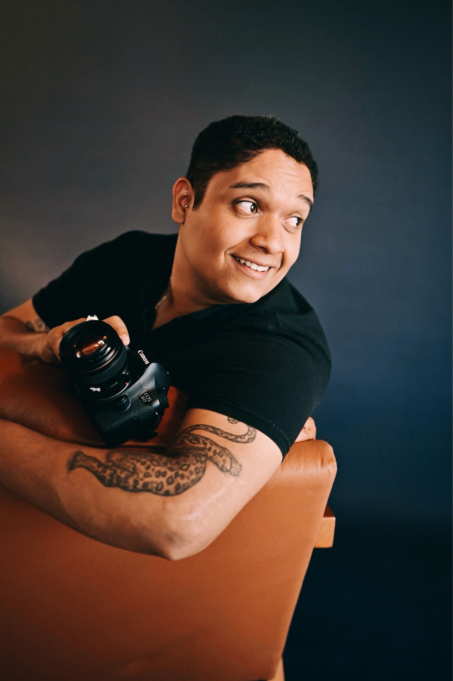 Smiling man with tattoos holding a camera while leaning over a brown chair against dark background.