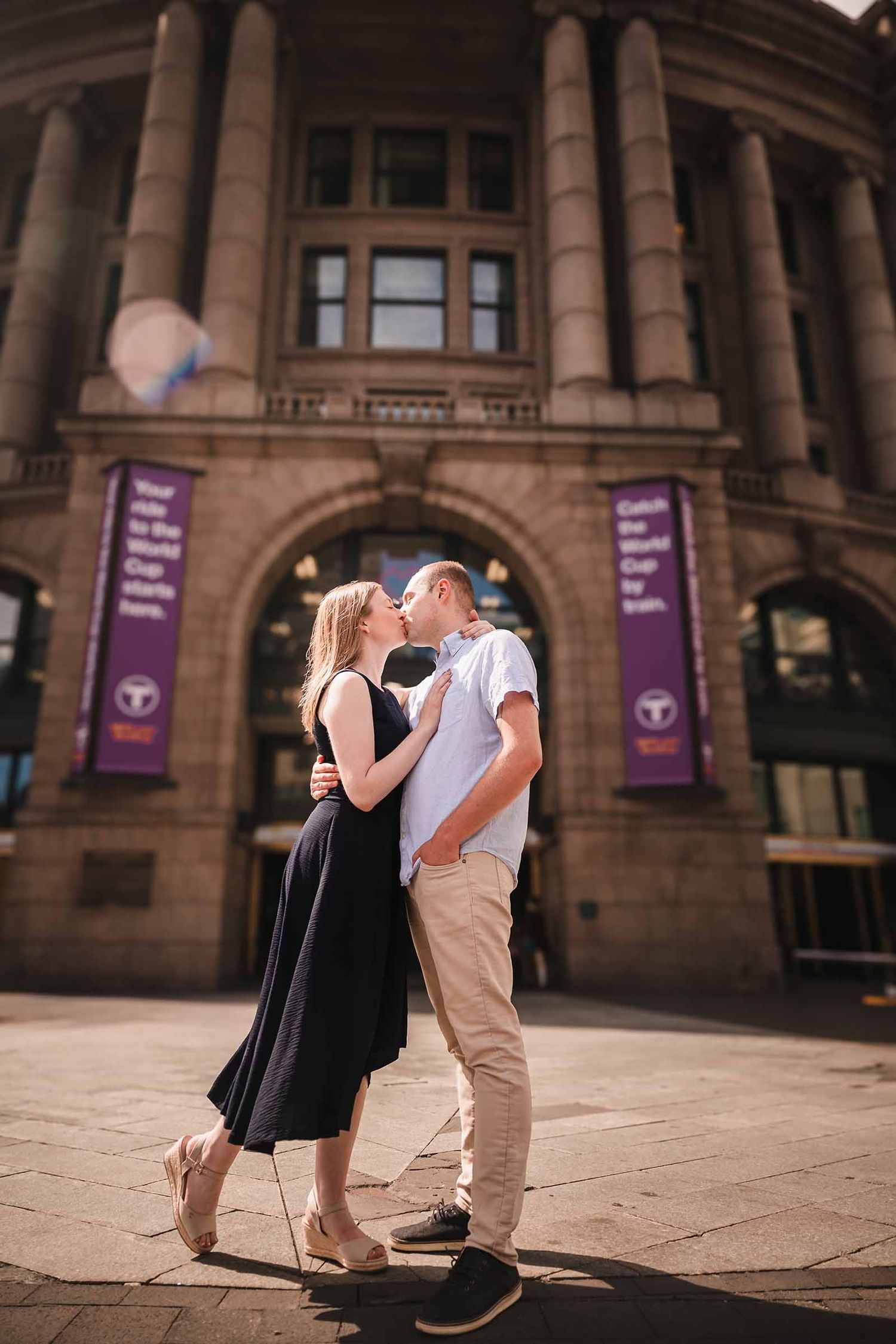 Engaged couple embracing outside South Station's clock tower with World Cup crowds and event barricades in the background