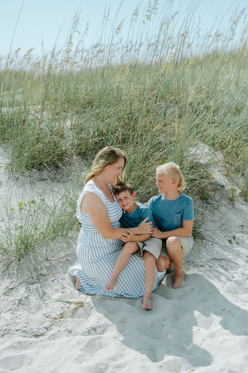 A woman in a striped dress sits on beach sand dunes with two young boys, smiling warmly together.
