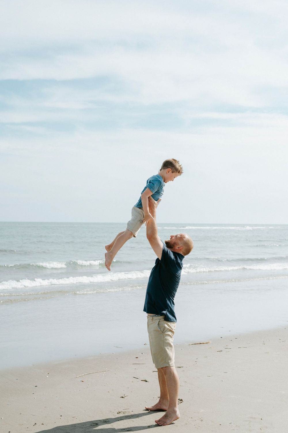 Father joyfully lifts young son high in the air on a sunny beach with ocean waves in the background.