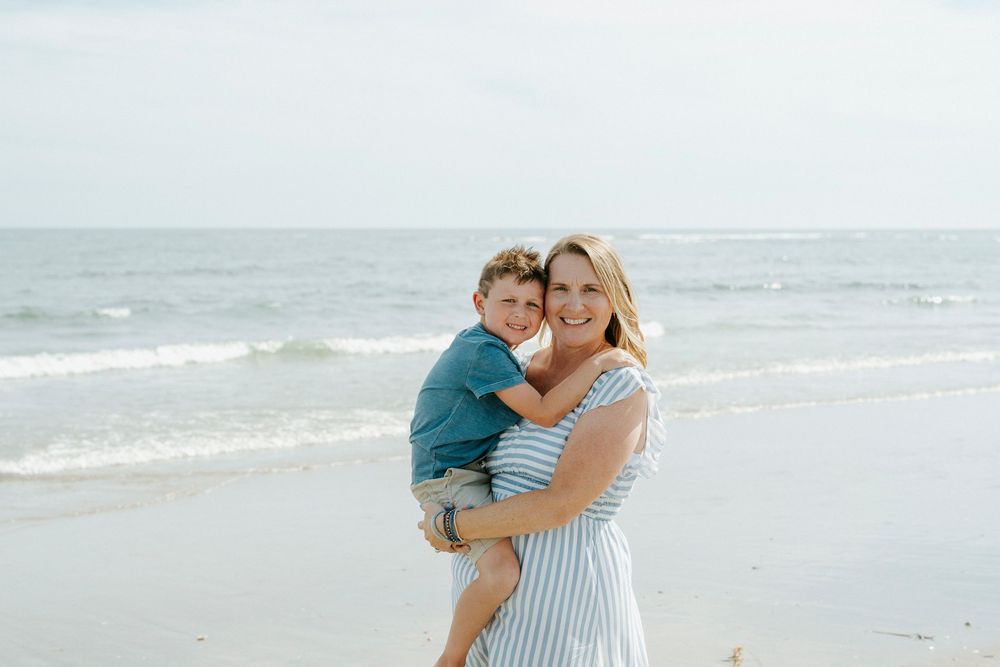 A smiling woman in a striped dress holds a young boy on a sunny beach with ocean waves in the background.
