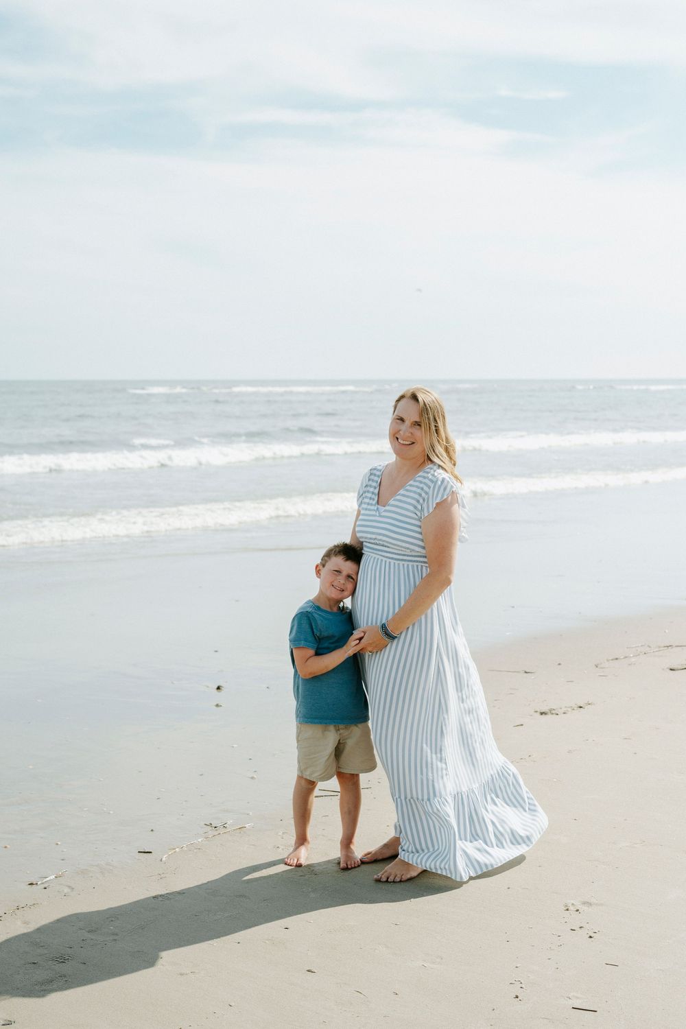Pregnant mother in striped maxi dress poses with young son hugging her belly on a sandy beach.
