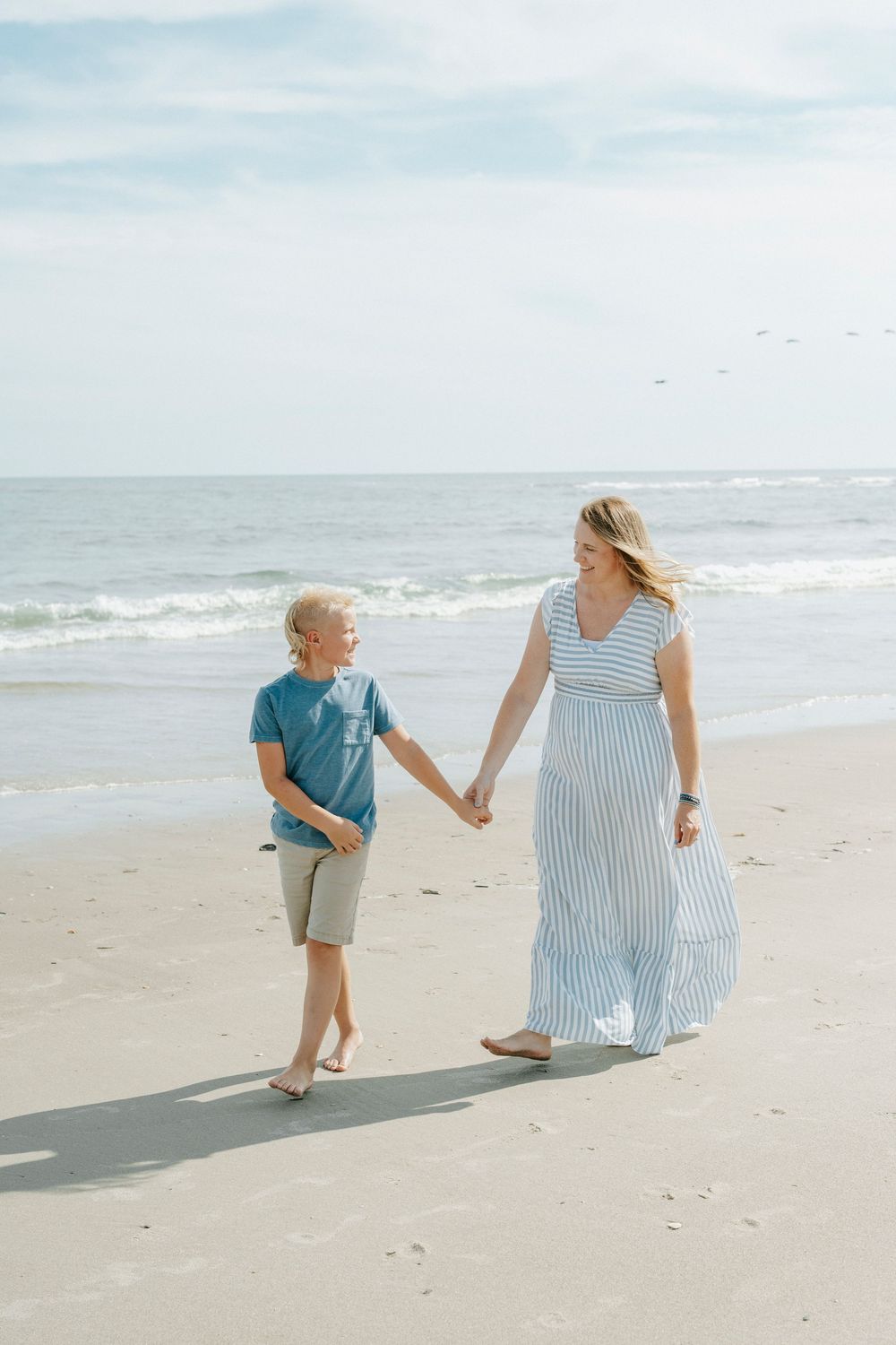 A woman in a striped maxi dress holds hands with a young boy on a sunny beach near the ocean.