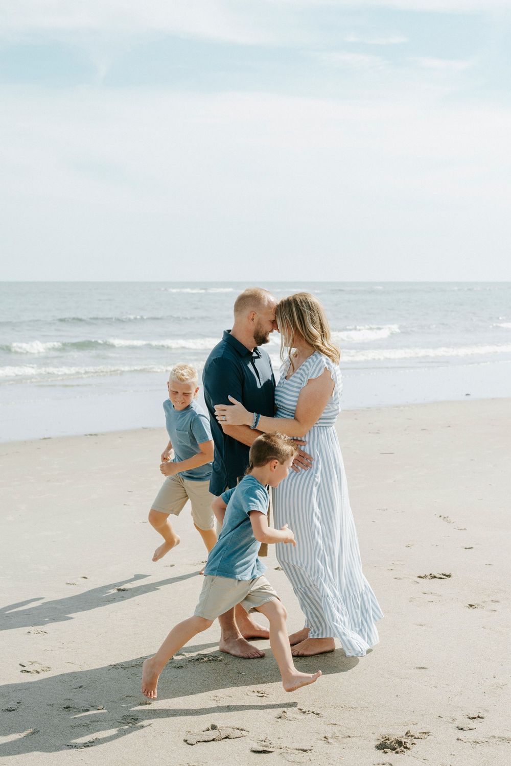 A family of four enjoys a beach day, with two young boys running while parents embrace near the ocean shoreline.