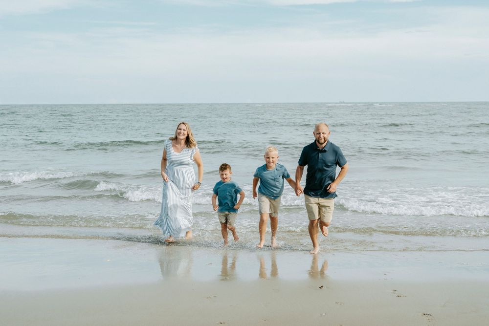 Family of four walking on beach shoreline, waves behind them, wearing casual summer clothes on a sunny day.