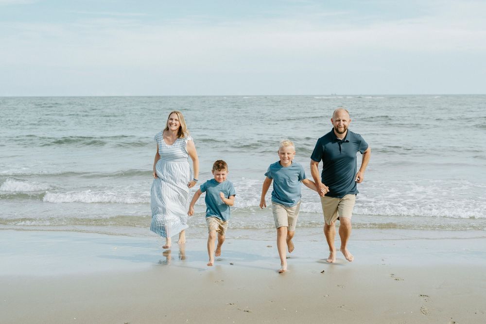Family of four running barefoot on a sandy beach with ocean waves in the background on a sunny day.