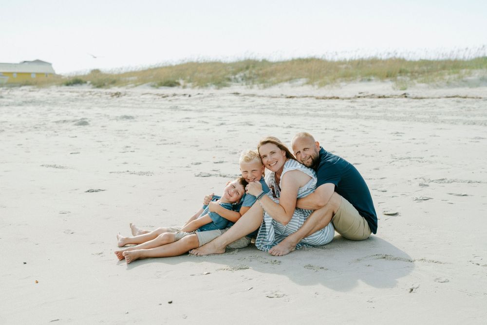 Family of four sitting together on a sandy beach, smiling and embracing in casual summer outfits.