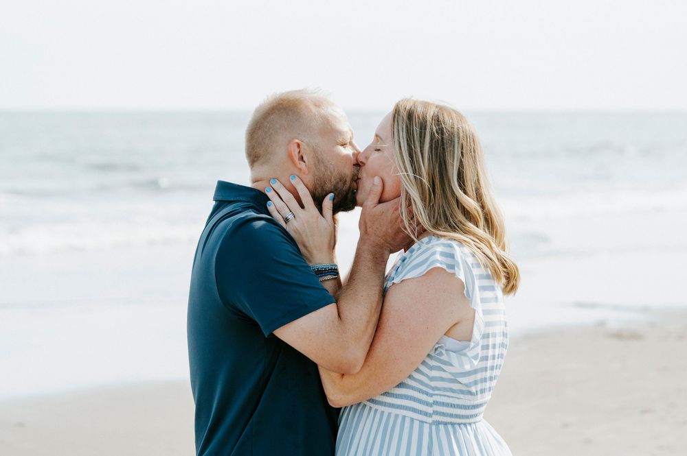 Couple sharing a romantic kiss on the beach with ocean waves in the background during an engagement photo session.