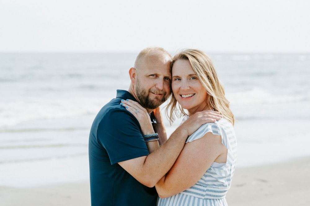 A smiling couple embraces affectionately on a beach with gentle ocean waves behind them.