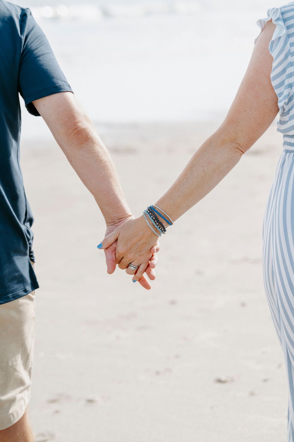 Couple holding hands on a sandy beach, woman wearing rings and blue bracelets, man in navy shirt and khaki shorts.