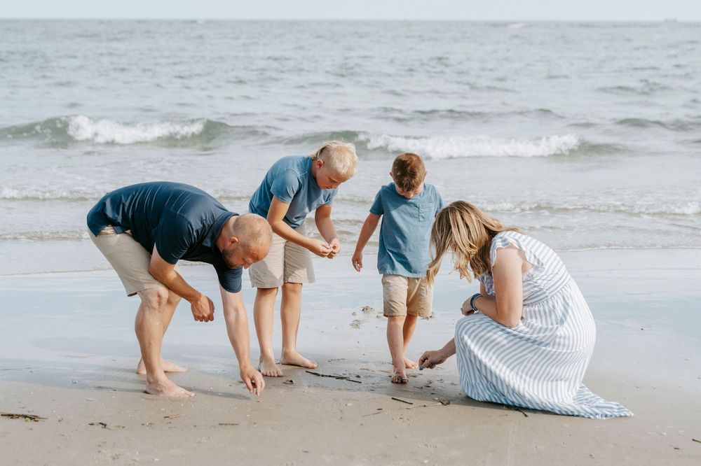 Family of four enjoying a beach day, bending down to explore shells and sand at the water's edge on a sunny day.